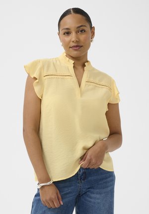 Femme aux cheveux tirés en arrière portant un chemisier jaune à manches courtes avec des volants et des détails en dentelle, associé à un jean bleu et des bijoux argentés.