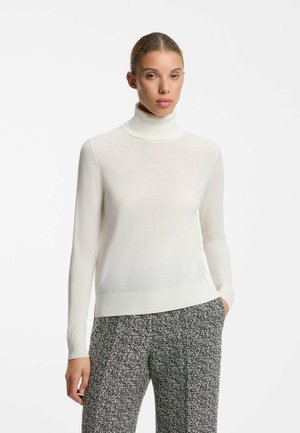 Woman standing wearing a white turtleneck sweater and black and white patterned pants, with one hand in her pocket against a white background.