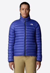 Giacca isolante blu con motivo a quadrati orizzontali, colletto alto e zip completa. Presenta il logo sul lato sinistro del petto e una texture di tessuto liscia.