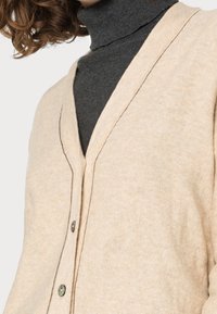 Beżowy kardigan z dekoltem w serek, teksturowaną tkaniną i widocznymi szwami. Posiada dwa guziki z przodu z detalem w kontrastowym kolorze.