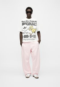 Άνδρας με μαύρα γυαλιά ηλίου, λευκό μπλουζάκι Adidas Originals με κίτρινο παπάκι, ανοιχτό ροζ παντελόνι και λευκά αθλητικά παπούτσια, στέκεται με τα χέρια στις τσέπες.