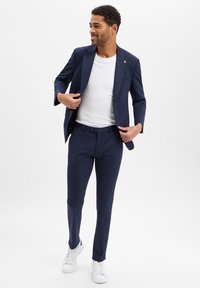 Marineblauer Anzugjacke und Hose kombiniert mit einem weißen T-Shirt und weißen Sneakers. Der Anzug hat eine schmal geschnittene Passform und einen glatten Stoff.