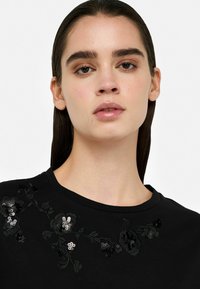 Schwarzes T-Shirt mit floralen Stickereien und Pailletten in Grautönen entlang des Ausschnitts. Glattes Material und klassischer Rundhalsausschnitt.