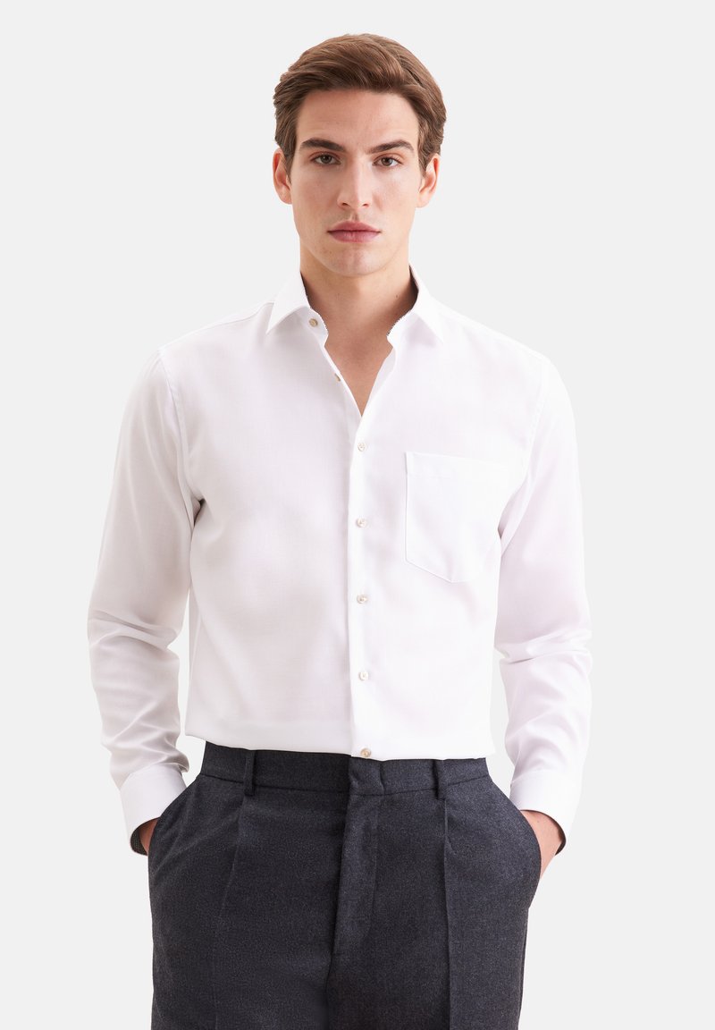 Jeune homme aux cheveux bruns porte une chemise blanche à manches longues boutonnée et un pantalon gris foncé, debout, les mains dans les poches.