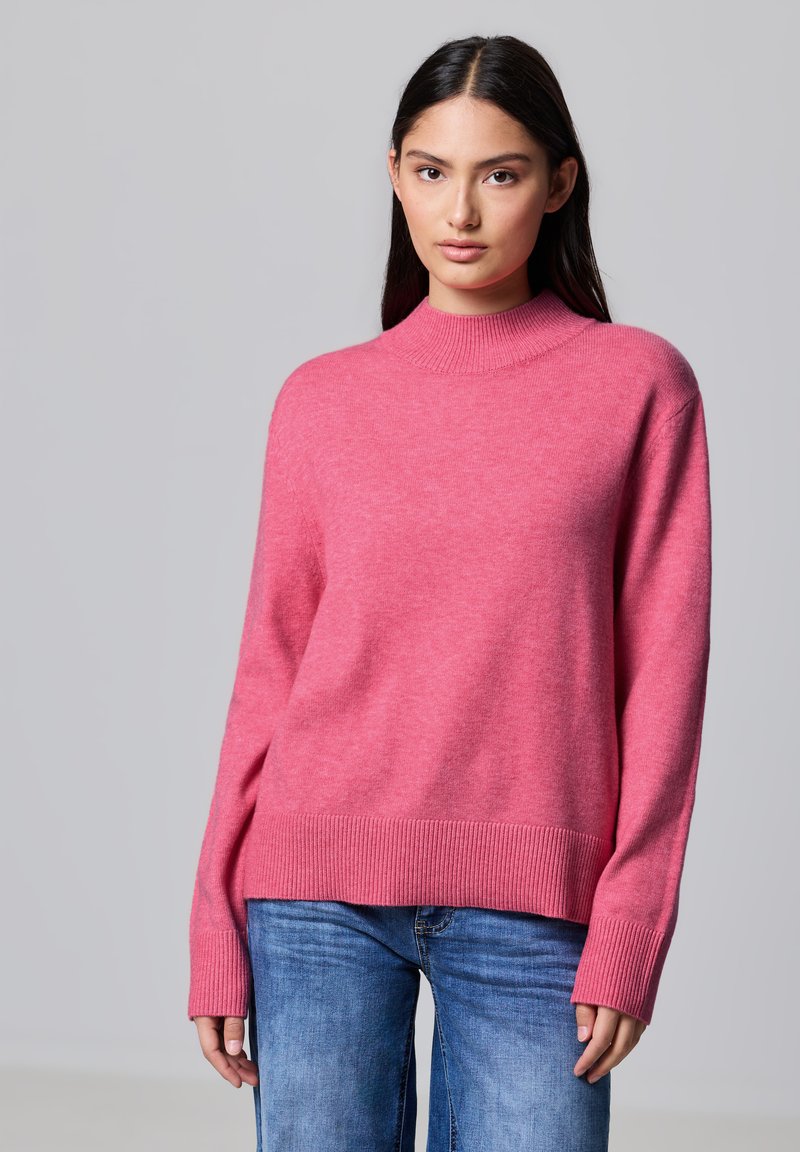Maglione rosa brillante con un colletto alto, polsini e orlo a coste. Realizzato in un materiale morbido, abbinato a jeans di denim blu.