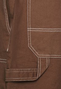 Tela de algodón marrón con textura diagonal, que presenta costuras blancas en contraste y un bolsillo con bordes angulados. Diseño simple y funcional.