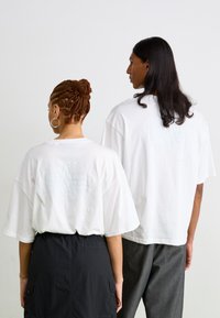 Hvit oversized t-skjorter med et teksturert logo på baksiden, med l�øs passform og korte ermer, båret av to personer bakfra.