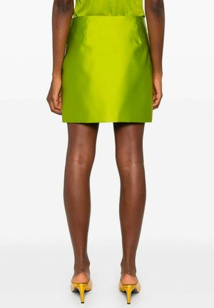 Kvinde iført en limegrøn mininederdel og matchende top, stående barfodet i gule kitten heels, vist bagfra mod en ensfarvet baggrund.