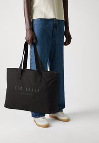 Black canvas tote bag with two sturdy handles, featuring "TED BAKER LONDON" logo in gray, rectangular shape, and a smooth texture.