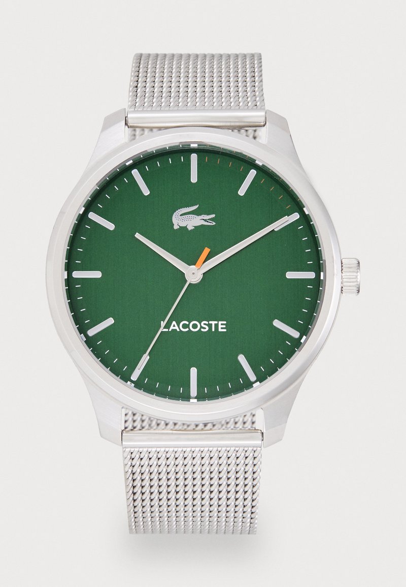 Round green dial watch with silver mesh band. Features white hour markers, orange second hand, and Lacoste logo. Smooth metal finish.