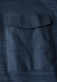 Námorníkovská texturovaná látka se splývací kapsou s chlopní. Kapsa má rovné okraje a viditelné prošívání. Detailní pohled.