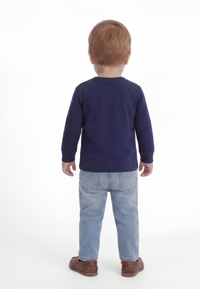 Niño pequeño pelirrojo de espaldas, vistiendo camiseta de manga larga azul marino, pantalones vaqueros claros y zapatos marrones sobre fondo blanco.