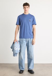 Zils kokvilnas T-krekls ar krūšu kabatu un drukātu logo, kopā ar gaiši ziliem plašiem džinsiem. Individuāls cilvēks tur zilu džemperi.