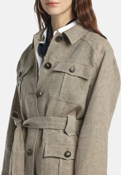 Nő bézs színű, öves lenvászon dzsekit visel nagy, gombos zsebekkel és gallérral, fehér inggel és sötét mintás sállal párosítva.