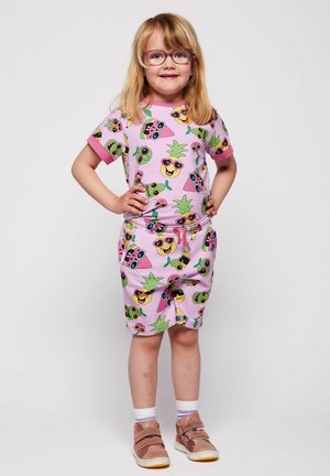 Jeune fille portant un t-shirt et un short roses assortis avec des motifs colorés de fruits souriants, des lunettes et des baskets roses, debout les mains sur les hanches.