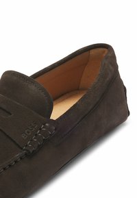 Mocasines de ante marrón oscuro con un acabado texturizado, que presentan un acento trenzado y una sutil marca en el lado. Forro interior suave visible.