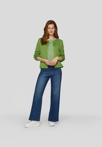 Grüner strukturierter Cardigan über einem weiß-gestreiften Shirt in Grün, kombiniert mit dunkelblauen weiten Jeans und weißen Sneakers.