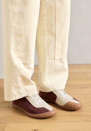 Αθλητικά παπούτσια από καμβά και σουέτ σε μπορντό και μπεζ, με υφασμάτινη λάστιχη σόλα και λευκές κορδόνες, συνδυασμένα με κρέμα χρώματος φαρδιά παντελόνια.