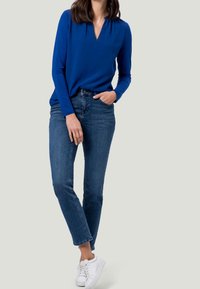 Donna con una maglietta blu a maniche lunghe, jeans blu a vita alta e sneakers bianche, in piedi con una mano in tasca e l'altra rilassata.