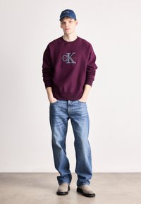 Maroonská mikina s logem "CK", sladěná se světle modrými džíny a šedými nazouvacími botami. Model má na sobě námořnickou kšiltovku.