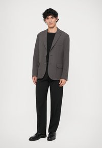 Blazer gris avec deux boutons et une poche poitrine, associé à un pantalon et une chemise noirs. Tissu doux et coupe ajustée. Des chaussures noires complètent le look.