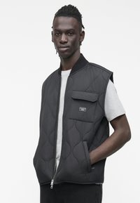 Μαύρο καπιτονέ γιλέκο με φερμουάρ μπροστά, ριμπ γιακά και τσέπη στο στήθος. Φορεμένο πάνω από γκρίζο πουκάμισο με υφή.