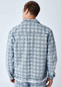 Veste en denim bleu délavé avec une texture à motifs présentant des formes en diamant, un col classique et une fermeture à boutons. La vue de dos montre des détails de couture.