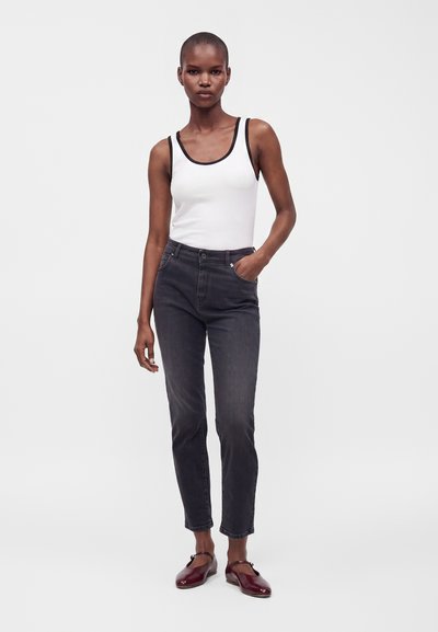 Femme portant un débardeur blanc avec une bordure noire, un jean foncé taille haute et des chaussures Mary Jane bordeaux, debout avec une main dans la poche.