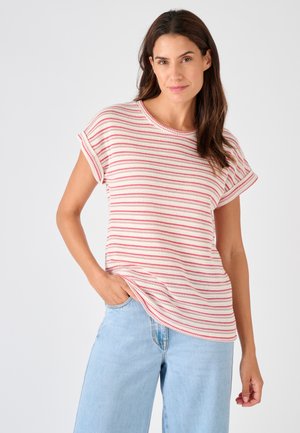 Donna con capelli castani lunghi che indossa una maglietta a maniche corte bianca con strisce rosa e jeans a vita alta azzurri, con una mano in tasca.