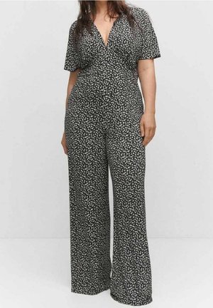 Frau in einem schwarzen Overall mit kurzen Ärmeln und kleinem weißem Blumenmuster, stehend vor einem schlichten hellen Hintergrund.
