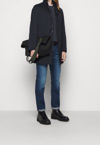 Tmavý, lehký kabát s vysokým límcem, zapínání na knoflíky, zkombinovaný se šedým svetrem a modrými džíny. Černé šněrovací boty a zelená kabelka na rameno.