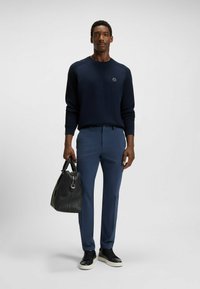 Granatowy sweter dziergany, niebieskie spodnie i czarne trampki. Model trzyma fakturowaną torbę z czarnej skóry. Prosty, dopasowany krój i swobodny styl.