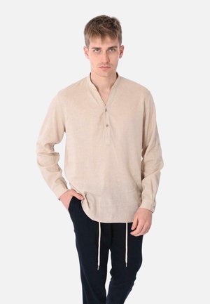 Camicia beige a maniche lunghe con colletto alla cinese, patta con bottoni frontale e spacchi laterali. Abbinata a pantaloni scuri con vita regolabile.