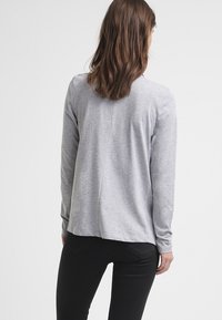 Graues Langarmoberteil mit lockerem Schnitt, ausgestattet mit einem Rückennähtedetail und einer weichen Stofftextur. Kombiniert mit schwarzen Hosen, von hinten betrachtet.