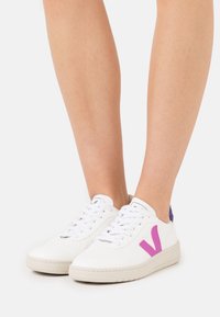 Zapatillas de cuero blancas con logo 'V' rosa, punta perforada, cordones blancos y acento púrpura en el talón. Estilo bajo con suela texturizada.