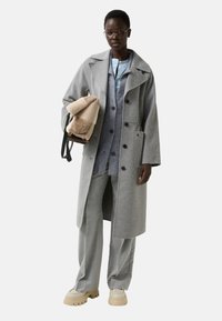 Tall person wearing oversized grey coat, light blue shirt, matching grey trousers, chunky beige shoes, holding beige and tan shearling bag.