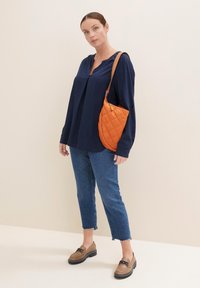Blusa azul marino con escote en V, bolso acolchado naranja claro en el hombro, jeans cortos azul claro y zapatos marrones con una correa decorativa.