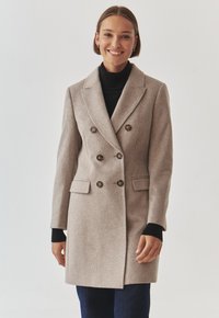 Abrigo beige de doble botonadura hecho de lana, con cuello de muesca, botones decorativos y bolsillos laterales. Llevado sobre un suéter de cuello alto negro.