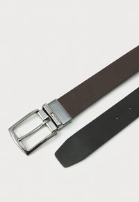 Cinturón de cuero marrón con una hebilla rectangular de plata, que presenta un pequeño logo. La correa tiene agujeros para ajustarse y un lado inferior negro liso.
