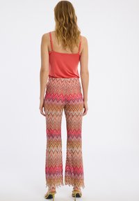 Uitlopende broek van een gebreide stof met een patroon van zigzagstrepen in roze, rood, oranje en bruin met een franjerand; gecombineerd met een koraalkleurige top.