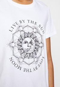 Camiseta blanca con un gráfico central que presenta un diseño de sol y luna, rodeado de patrones geométricos y el texto "VIVE POR EL SOL, AMA POR LA LUNA."