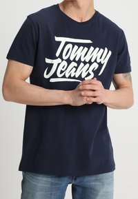 Mörkblå bomull t-shirt med rund halsringning som har en stor vit "Tommy Jeans"-logotyp i handskriven stil över bröstet.
