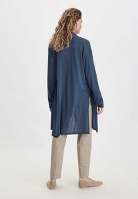 Langer navyfarbener Cardigan aus leichtem Stoff, mit lockerer Passform, Seitenschlitzen und überschnittenen Schultern, kombiniert mit beigen Hosen.