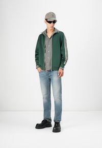Veste zippée verte avec des rayures blanches, chemise à carreaux, jeans bleu clair et chaussures noires chunky. Une casquette et des lunettes de soleil complètent le look.