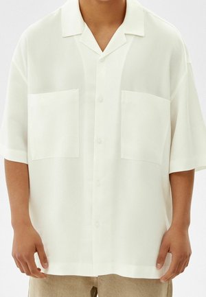 Persoon draagt een losse, korte mouwen witte blouse met twee voorzakken en beige broek tegen een effen achtergrond.