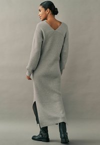 Robe maxi en tricot côtelé gris avec un large col en V dans le dos, des fentes sur les côtés et des manches longues. Associée à des bottes noires à bout carré.