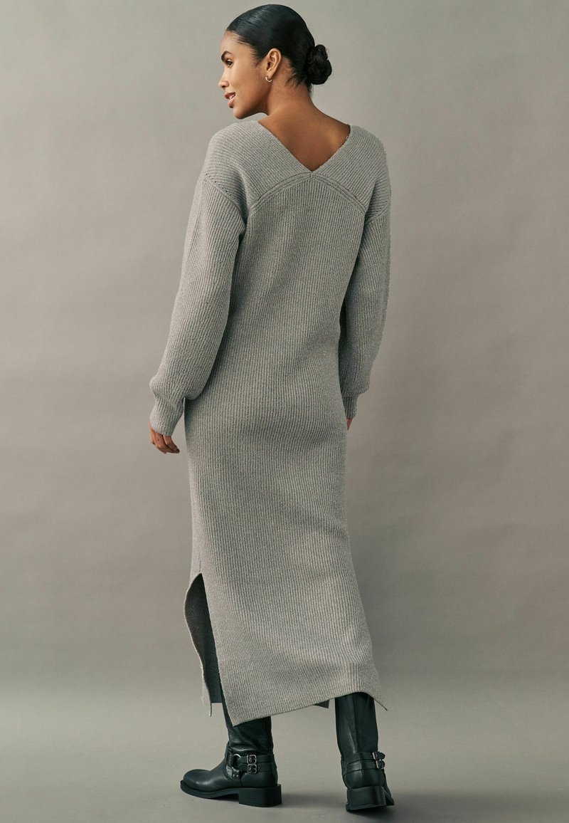 Robe maxi en tricot côtelé gris avec un large col en V dans le dos, des fentes sur les côtés et des manches longues. Associée à des bottes noires à bout carré.