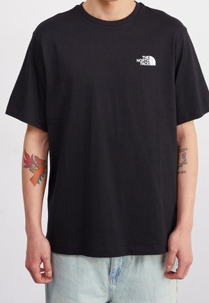 Henkilö pukeutunut mustaan The North Face -T-paitaan, jossa on lyhyet hihat ja tatuoinnit molemmissa kyynärvarsissa, yhdistettynä vaaleansinisiin farkkuihin.