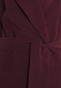 Abrigo envolvente burdeos con cinturón, hecho de tela suave. Presenta un cuello de chal y un detalle de costura visible, mostrando una superficie lisa y texturizada.
