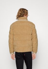 Tan nepbont pufferjack met horizontale stiksels, hoge kraag en elastische manchetten. Zwarte broek zichtbaar bij de taille. Zachte textuur.